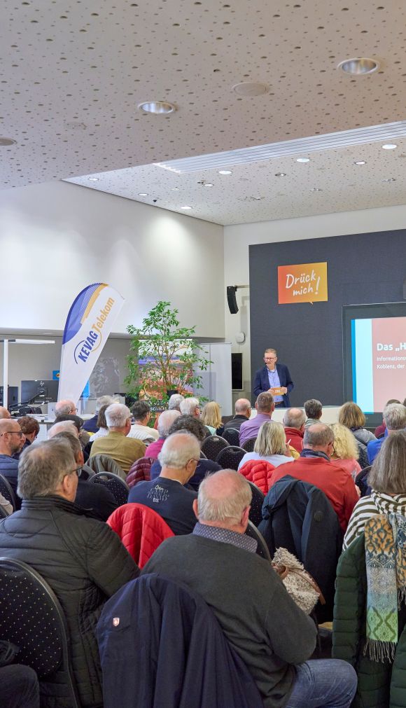 Ein Publikum sitzt in einem Saal. Vor ihnen steht ein professionell gekleideter Mann und präsentiert..