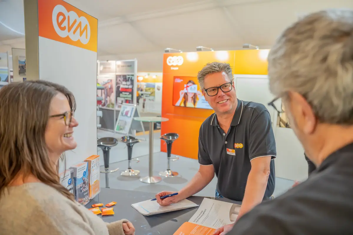 Drei Personen stehen an einem Stand. Die brünette Frau und der Mann mit den weißen Haaren befinden sich am Bildrand und man kann nur die Hinterköpfe sehen. In der Mitte steht ein Mann der den Mann mit den weißen Haaren anlächelt.