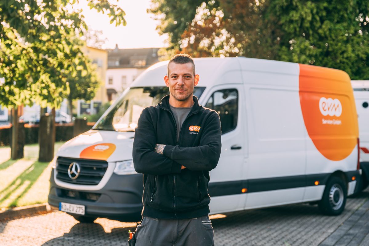 Ein Mann steht mit verschränkten Armen und lächelt vor einem weißen Mercedes-Transporter mit oranger Beschriftung "EVM Service GmbH". Der Hintergrund zeigt eine sonnige, baumbestandene Straße.