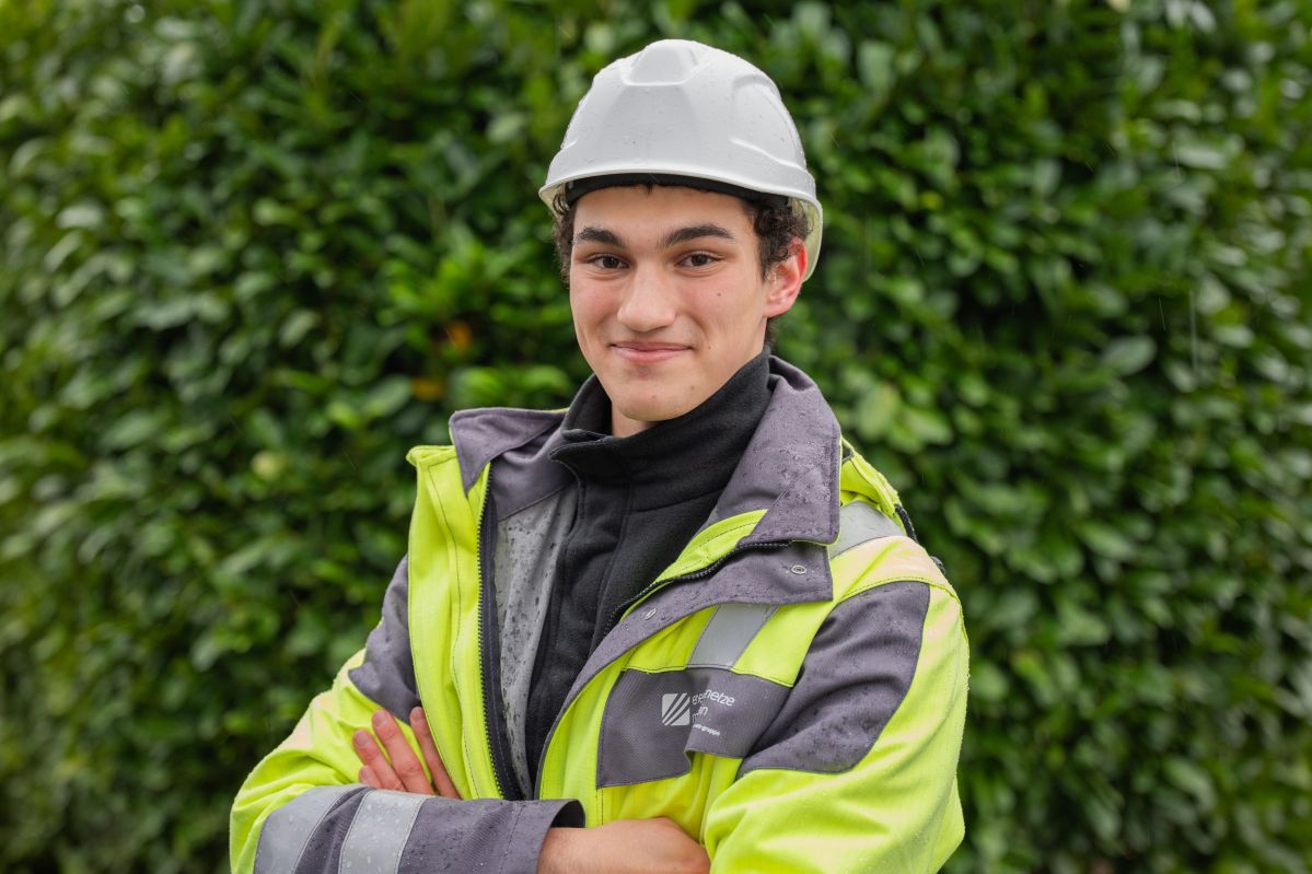 Präzisionsarbeit: Einblicke in die Ausbildung zum Geomatiker Ein lächelnder junger Mann mit weißem Schutzhelm und gelber Warnjacke steht mit verschränkten Armen; im Hintergrund ein dichter, grüner Busch.