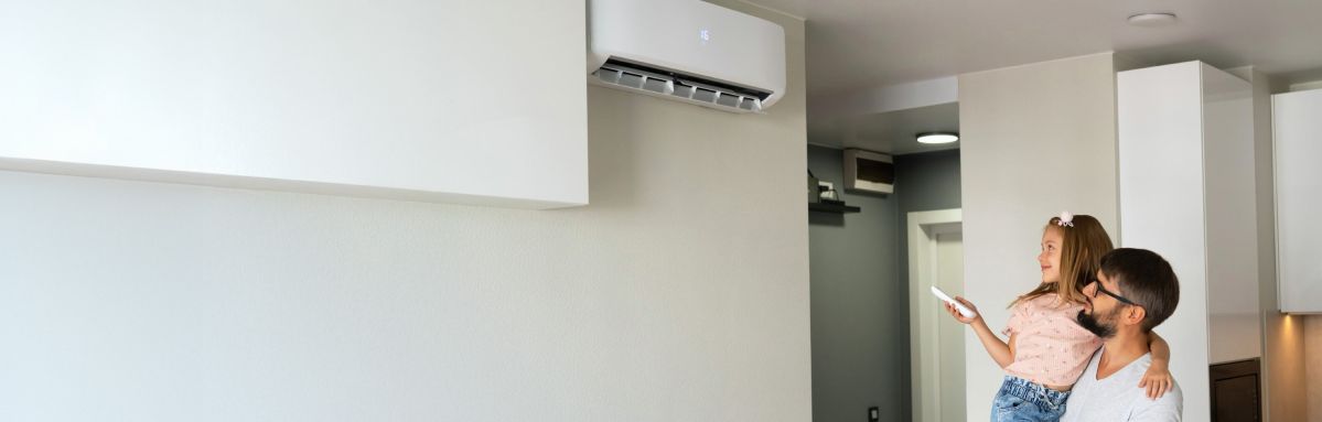 Ein Mann hält ein kleines Mädchen im Arm. Sie stehen in einem Wohnzimmer und schauen auf eine Klimaanlage, die an der Wand montiert ist. Das Mädchen hält eine Fernbedienung und zeigt mit dieser auf die Klimaanlage.