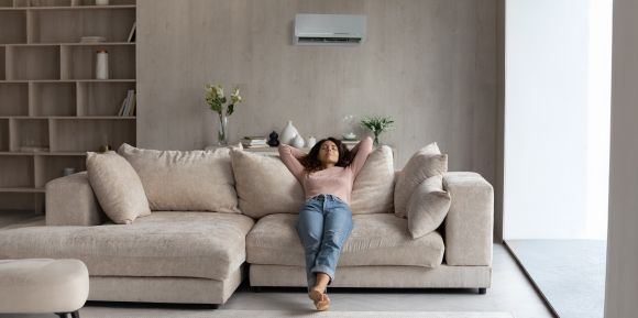 Eine Frau sitzt lässig auf einem beigen Sofa und hat ihre Arme hinter dem Kopf verschränkt. Über ihr befindet sich eine Klimaanlage.