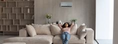 Eine Frau sitzt lässig auf einem beigen Sofa und hat ihre Arme hinter dem Kopf verschränkt. Über ihr befindet sich eine Klimaanlage.