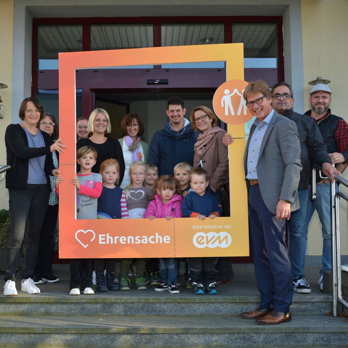 Mehrere Erwachsene und Kinder lächeln freundlich und halten einen orangenen Rahmen mit der Aufschrift "Ehrensache".
