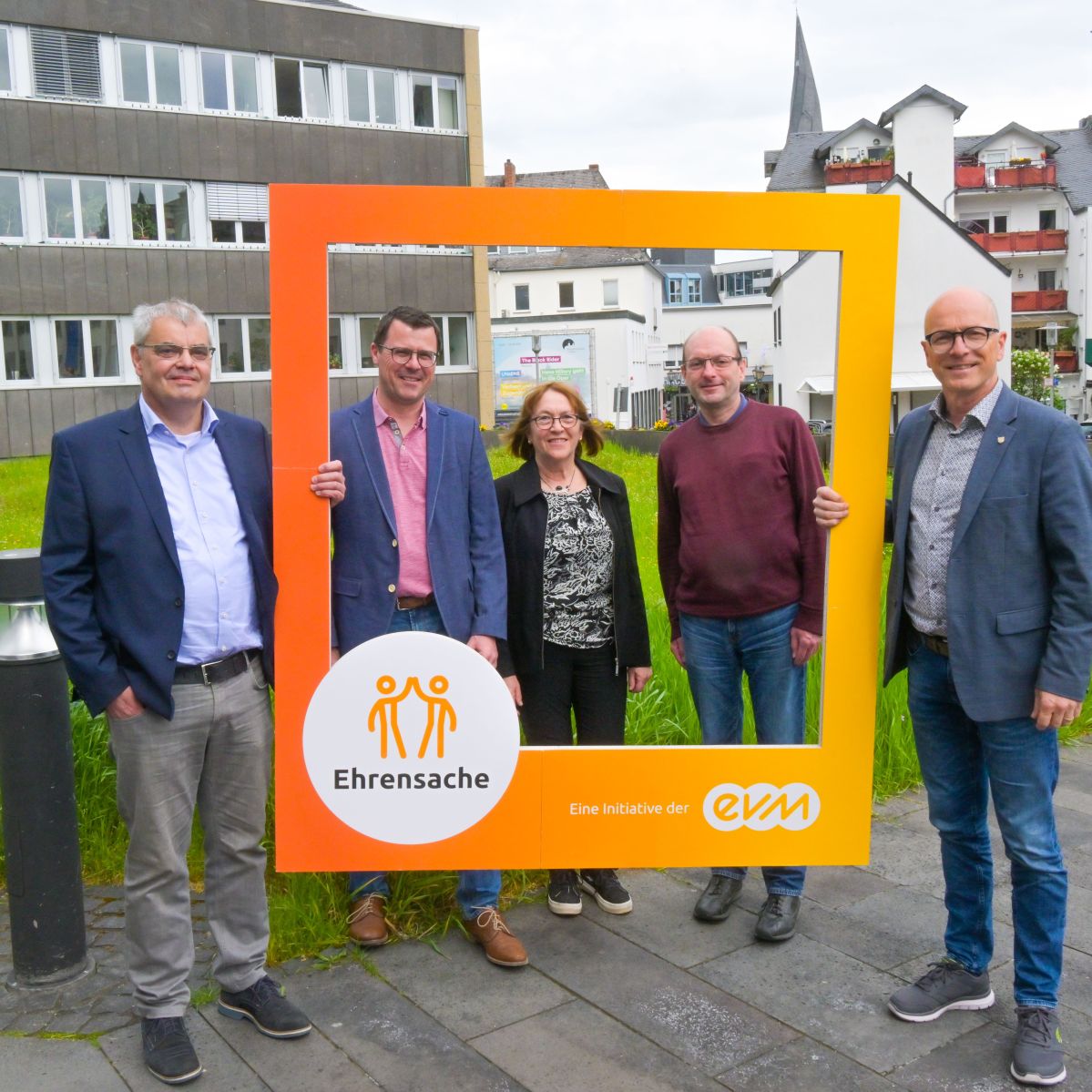 Vier Männer und eine Frau lächeln freundlich und halten einen orangenen Rahmen mit der Aufschrift "Ehrensache".