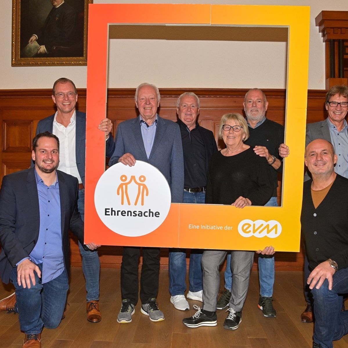 Sieben Männer und eine Frau lächeln freundlich und halten einen orangenen Rahmen mit der Aufschrift "Ehrensache".