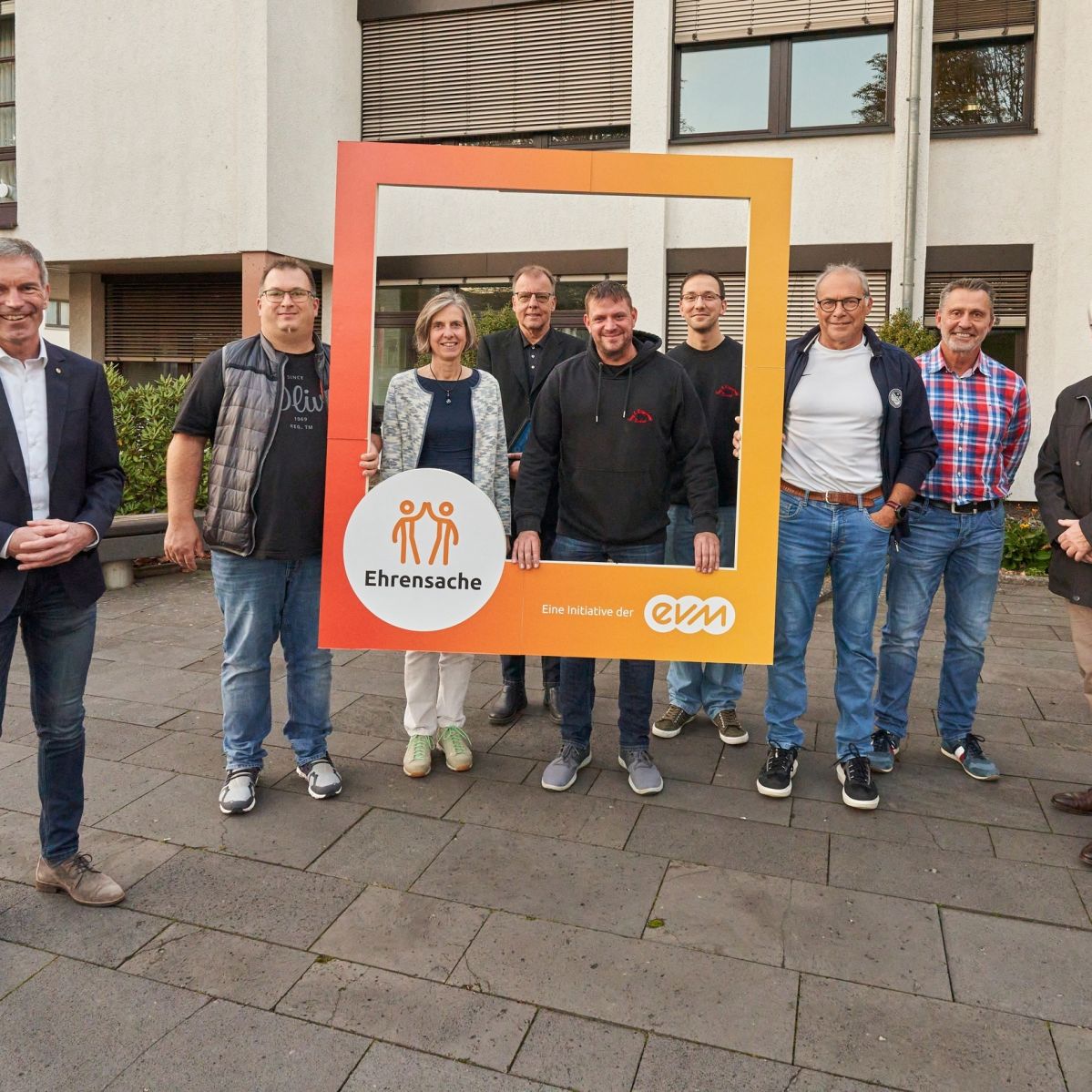 Acht Männer und eine Frau lächeln freundlich und halten einen orangenen Rahmen mit der Aufschrift "Ehrensache". 