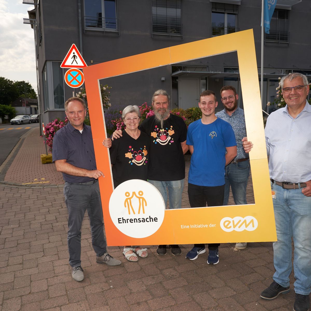 5 Männer und eine Frau lächeln freundlich und halten einen orangenen Rahmen mit der Aufschrift "Ehrensache".