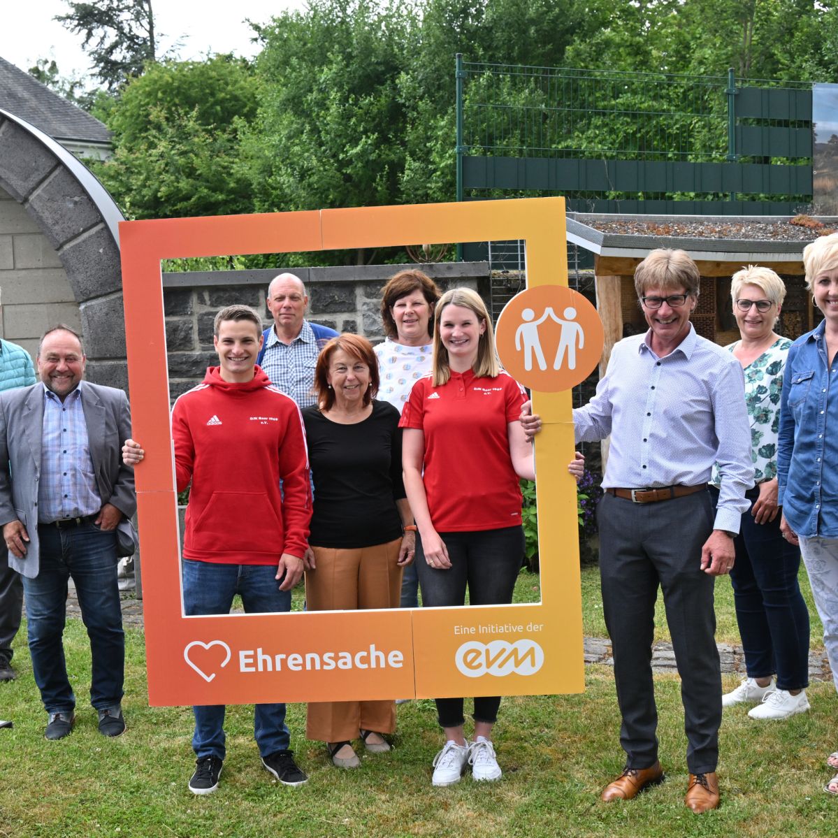 Fünf Männer und fünf Frauen lächeln freundlich und halten einen orangenen Rahmen mit der Aufschrift "Ehrensache".