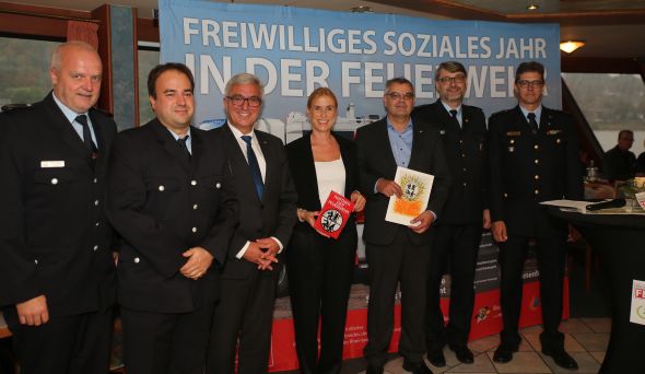 Sechs Männer und eine Frau sind professionell gekleidet und lächeln freundlich. Sie stehen vor einem großen Plakat mit Aufschrift "Freiwilliges soziales Jahr in der Feuerwehr" und halten Zertifikate.