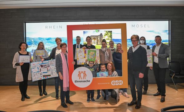 Eine Gruppe von Menschen, darunter zwei Kinder, hält bunte Zeichnungen und einen großen, rot-orangefarbenen Bilderrahmen mit der Aufschrift „Ehrensache, Eine Initiative der evm“. Im Hintergrund Landschaftsbilder.