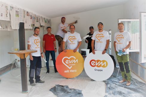 Sieben Männer mit "evm mit Herz" Tshirts stehe in einem Raum und haben Malwerkzeug in der Hand. Sie halten 2 kreisförmige Schilder mit der Aufschrift "evm mit Herz" und " evm Gruppe".