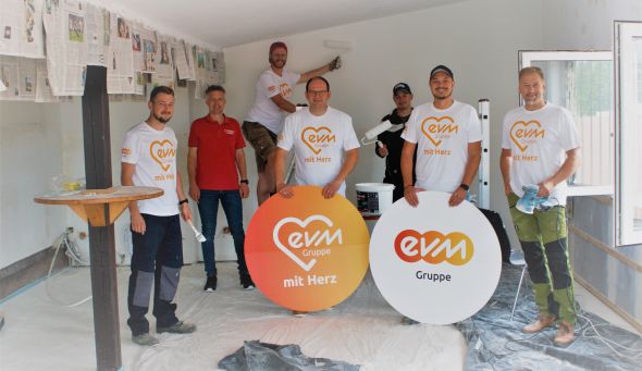 Sieben Männer mit "evm mit Herz" Tshirts stehe in einem Raum und haben Malwerkzeug in der Hand. Sie halten 2 kreisförmige Schilder mit der Aufschrift "evm mit Herz" und " evm Gruppe".