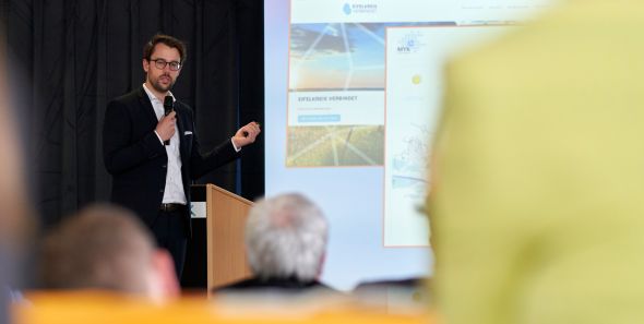 evm Smartmacher-Hausmesse: Innovative Technik für die Region Ein Mann steht mit Mikrofon an einem Rednerpult, präsentiert Folien unter dem Titel "Beispiele Rheinland-Pfalz" in einem Konferenzraum. Zuschauer sind im Vordergrund sichtbar.