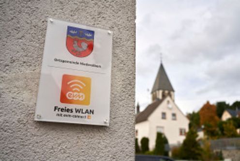 Ein Schild mit einem WLan Symbol und Aufschrift "evm Freies Wlan" hängt an einer Mauer. Im Hintergrund ist eine Kirche zu erkennen.