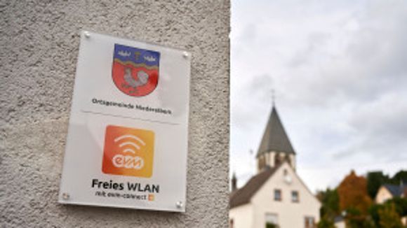 Ein Schild mit einem WLan Symbol und Aufschrift "evm Freies Wlan" hängt an einer Mauer. Im Hintergrund ist eine Kirche zu erkennen.