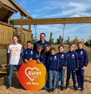 Mehrere Kinder in dunkelblauen Jacken und zwei Männer stehen auf einem Spielplatz und lächeln freundlich. Sie halten ein orangenes kreisförmiges Schild mit der Aufschrift "evm mit Herz".