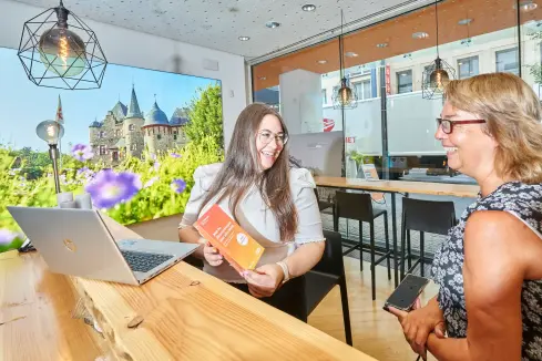 Zwei Frauen sitzen an einem Tisch in einem evm Kundenzentrum. Vor ihnen steht ein Laptop. Sie lächeln fröhlich und schauen auf einen Flyer.