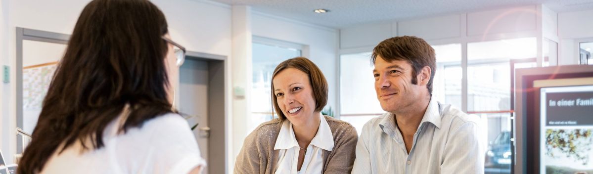 Beratungsgespräch Ein Paar sitzt an einem Schreibtisch in einer modernen Büroumgebung und bespricht Dokumente mit einer Beraterin. Ein Computerbildschirm und Büromaterialien sind im Hintergrund sichtbar.