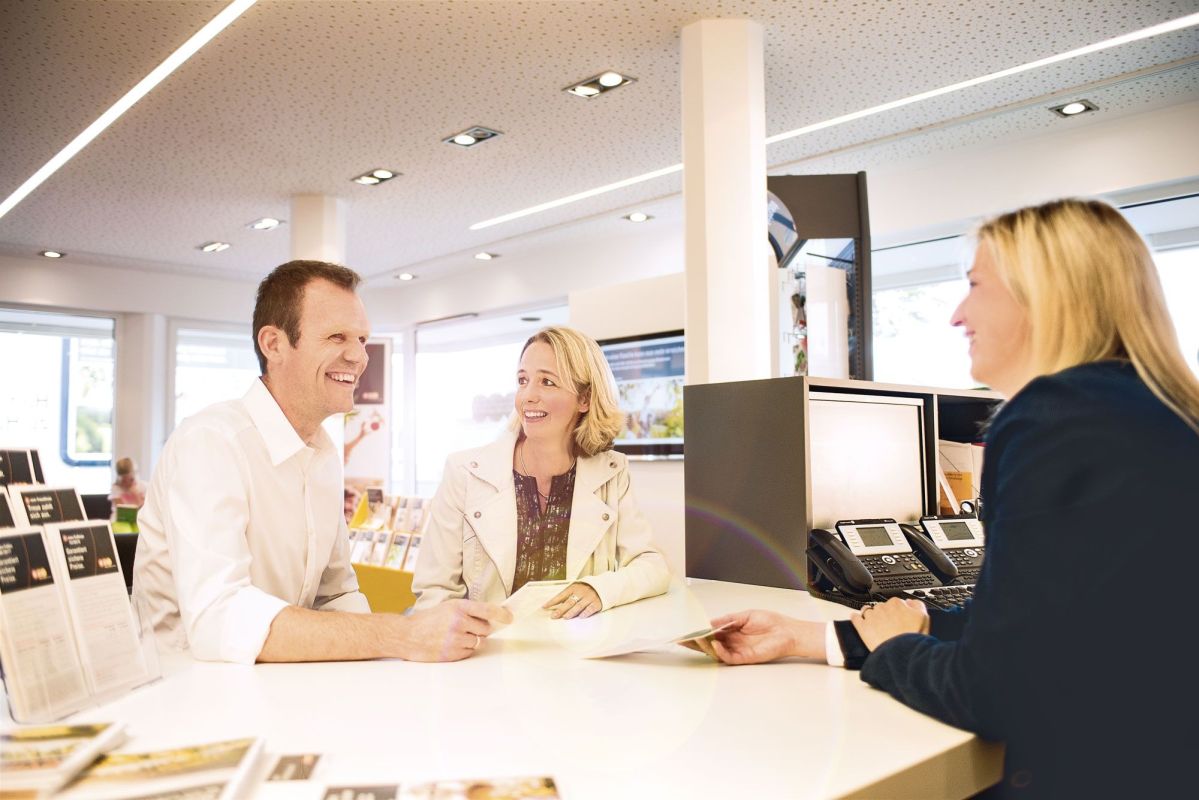 Ein Mann und eine Frau sitzen lächelnd an einem Schalter, während eine Frau ihnen gegenüber Informationen gibt; im Hintergrund ein modernes Büro mit Broschüren und Telefonen.