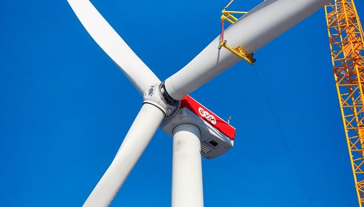 Eine große Windturbine rotiert gegen einen klaren blauen Himmel. Ein gelber Kran ist rechts sichtbar