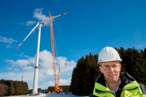 Ein Mann mit gelber Schutzjacke und einem Schutzhelm steht vor einem Windrad neben dem ein Kran steht.