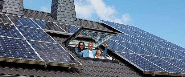 Drei Personen stehen lachend am offenen Dachfenster eines Hauses, das mit Solarpanels bedeckt ist. Der Himmel ist blau mit ein paar Wolken, was eine sonnige Umgebung suggeriert.