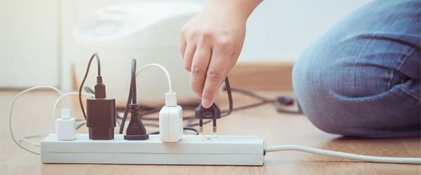 Eine Hand steckt einen Stecker in eine weiße Mehrfachsteckdose mit mehreren Kabeln; im Hintergrund ist ein weißes Haushaltsgerät auf Holzboden zu sehen.