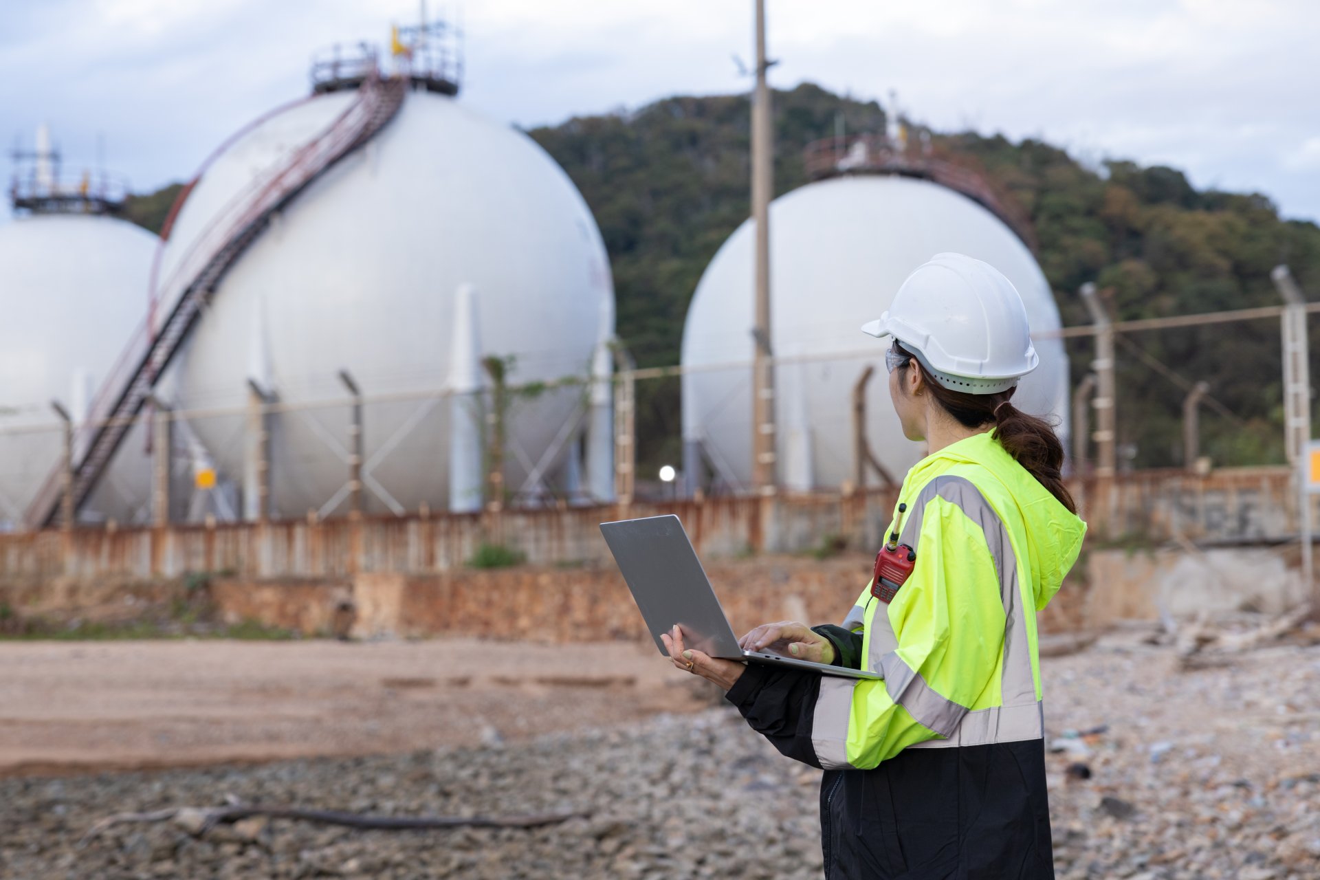 Gasnetz-Stilllegung? Warum die Versorgung sicher bleibt Das Bild zeigt eine Person in Sicherheitskleidung, die vor großen kugelförmigen Tanks steht. n den Händen hält die Person einen geöffneten Laptop und blickt in Richtung der Tanks, als würde sie Inspektionen oder Messungen durchführen.
