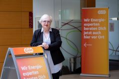 Eine ältere Frau mit weißen Haaren ist professionell gekleidet und steht vor einem evm Kundenzentrum und lächelt freundlich.