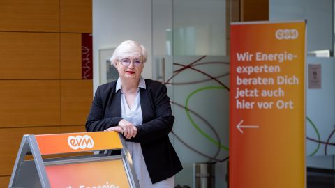 xxx Eine ältere Frau mit weißen Haaren ist professionell gekleidet und steht vor einem evm Kundenzentrum und lächelt freundlich.