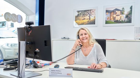 Eine Frau sitz an einem Schreibtisch und hält einen Telefonhörer an ihr Ohr. Vor ihr steht ein Bildschirm und eine Tastatur.