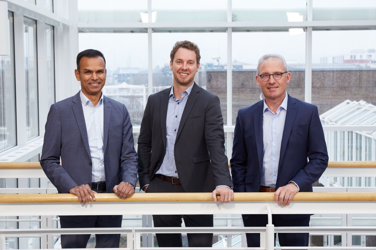 Drei professionell gekleidete Männer stehen auf einer Balustrade und lächeln freundlich.