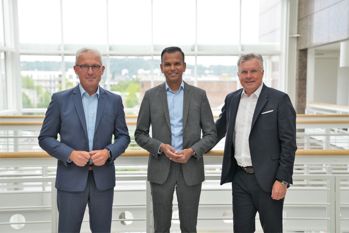 Drei professionell gekleidete Männer stehen vor einem urbanen Hintergrund und lächeln freundlich.
