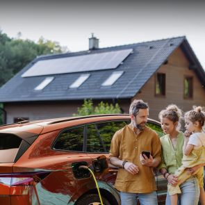 PV-Anlage und Wallbox: Das perfekte Duo Eine junge Familie steht an ihrem E-Auto. Im Hintergrund ist ein Haus mit Solaranlage auf dem Dach zu sehen