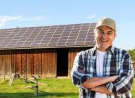 Solaranlage auf Scheune Eine Holzscheune mit Solarpaneelen auf dem Dach. Ein Mann mit gekreuzten Armen und Baseballkappe steht vor der Scheune, die von hellem Tageslicht umgeben ist.