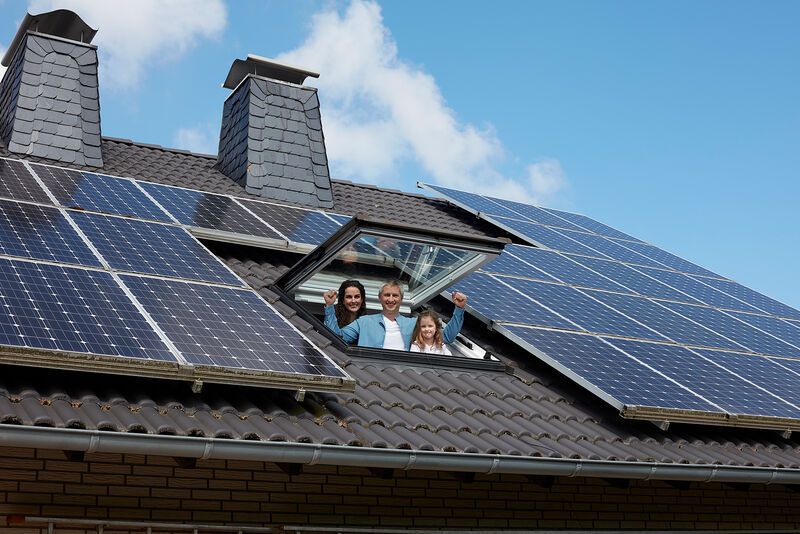 Drei Personen stehen lachend am offenen Dachfenster eines Hauses, das mit Solarpanels bedeckt ist. Der Himmel ist blau mit ein paar Wolken, was eine sonnige Umgebung suggeriert.