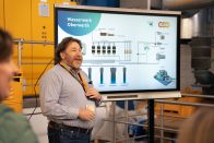 Ein Mann präsentiert lächelnd vor einem großen Bildschirm mit einer technischen Grafik und der Überschrift "Wasserwerk Oberwerth" in einem industriellen Raum mit gelben Maschinen im Hintergrund.