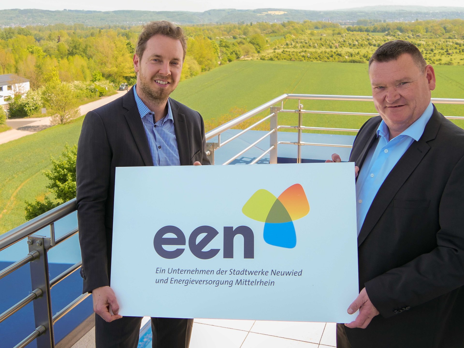 Zwei professionell gekleidete Männer lächeln freundlich in die Kamera und halten ein Schild mit der Aufschrift "een".