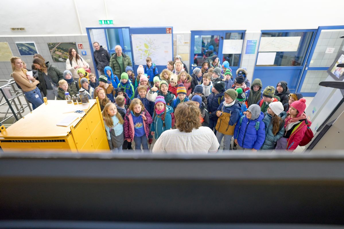 Ein Erwachsener erklärt einer Gruppe von Kindern etwas in einem Gebäude. Die Kinder tragen bunte Jacken und Mützen. Im Hintergrund sind Türen und an den Wänden hängen Infotafeln.