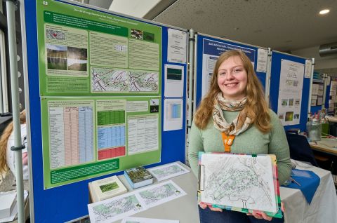 250210SDI1051 Ein Mädchen steht vor einer Wand mit Daten und hält ein Klemmbrett mit einer Karte und lächelt freundlich.