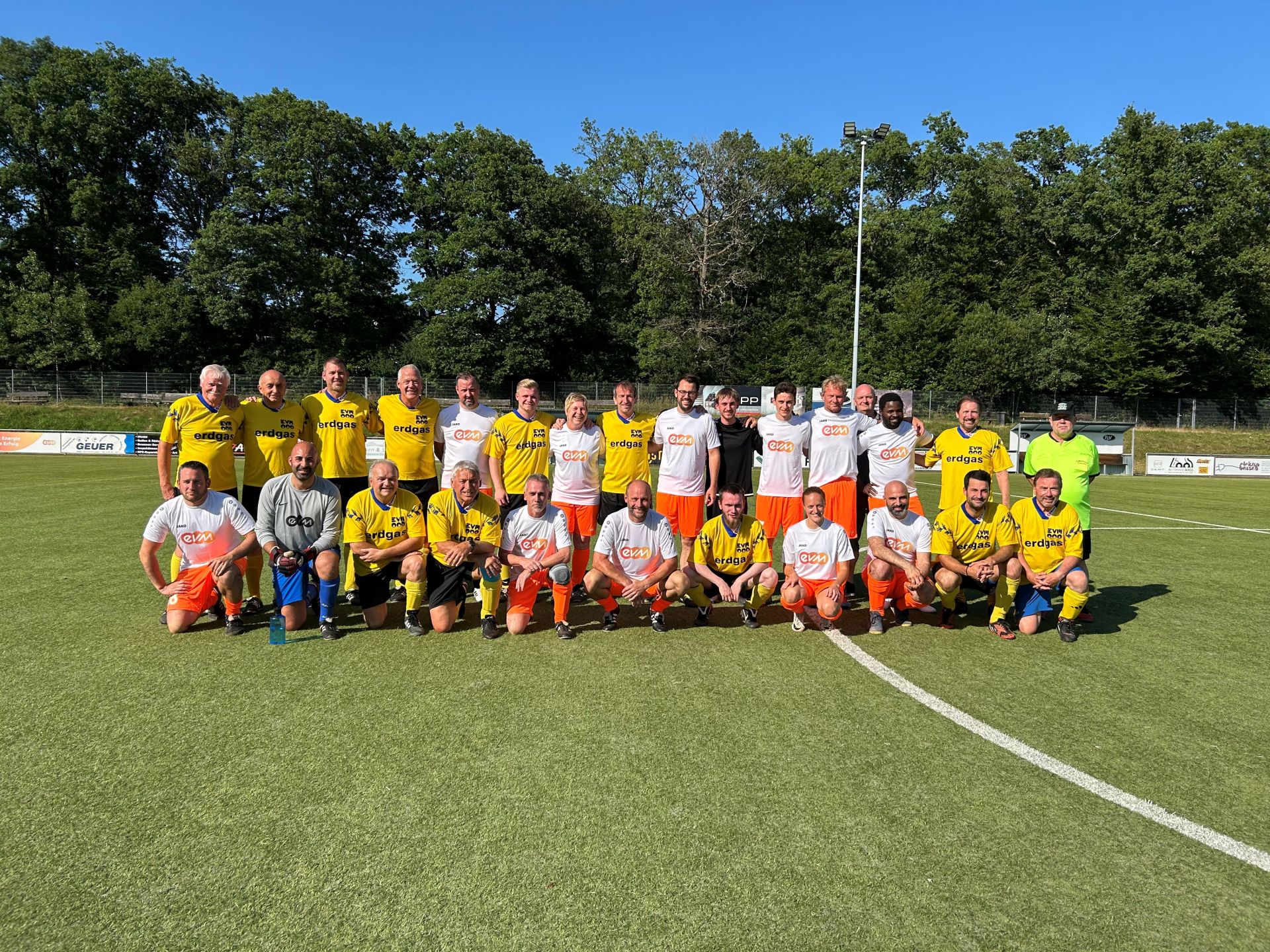 Zwei Fußballmannschaften stehen zusammen auf einem Fußballplatz und lächeln freundlich. Die eine Mannschaft trägt Gelb-Schwarze Trikots und die andere Weiß-Orangene Trikots mit evm-Logo