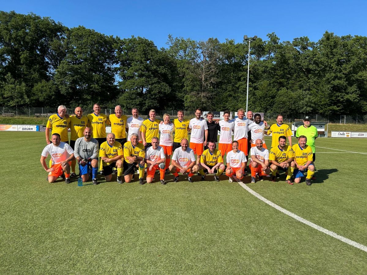 Zwei Fußballmannschaften stehen zusammen auf einem Fußballplatz und lächeln freundlich. Die eine Mannschaft trägt Gelb-Schwarze Trikots und die andere Weiß-Orangene Trikots mit evm-Logo