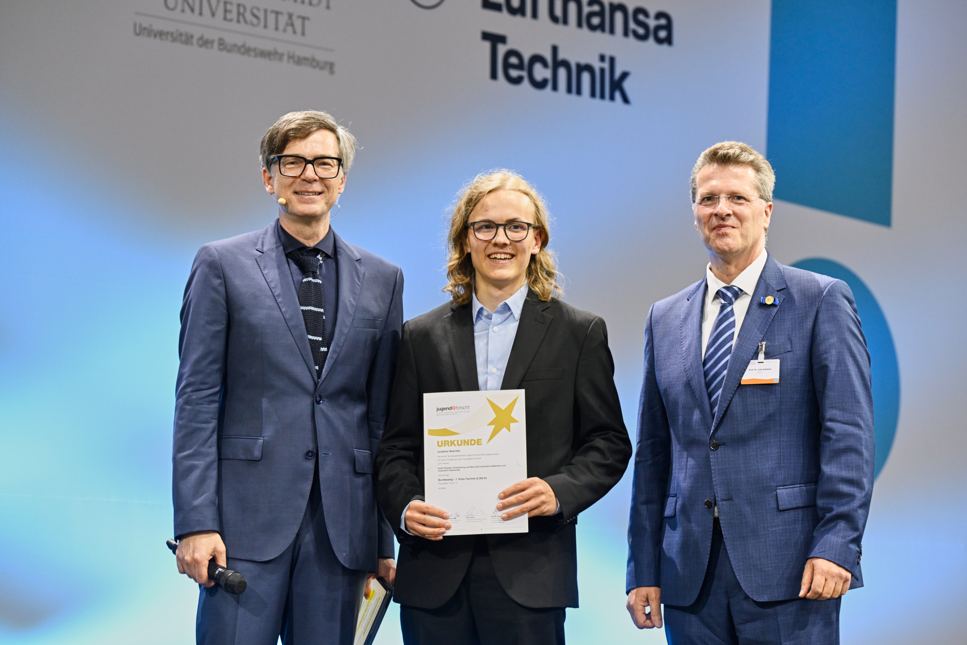 § Männer stehen auf einer Bühne und sind professionell gekleidet. Der junge Mann  der Mitte hält eine Urkunde.