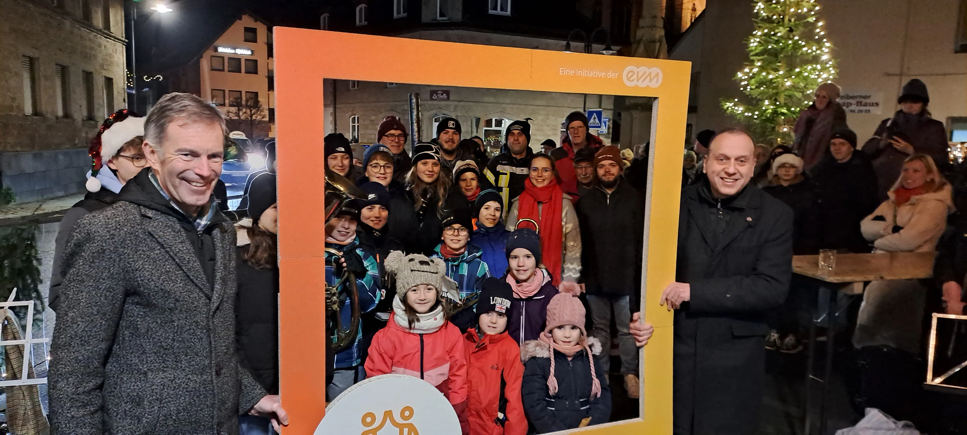 Eine Gruppe von Menschen posiert lachend mit einem großen, orangefarbenen Bilderrahmen auf einem nächtlichen Weihnachtsmarkt; hinter ihnen sind Lichter und ein geschmückter Weihnachtsbaum zu sehen.