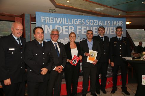 Sechs Männer und eine Frau sind professionell gekleidet und lächeln freundlich. Sie stehen vor einem großen Plakat mit Aufschrift "Freiwilliges soziales Jahr in der Feuerwehr" und halten Zertifikate.