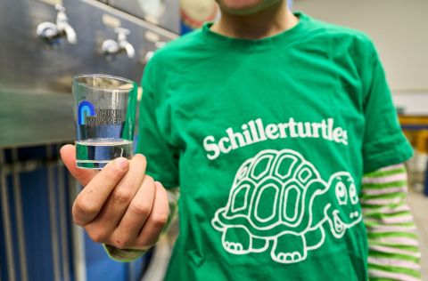 Ein Mann mit einem grünen T-shirt hält ein Glas mit der Aufschrift "Trinkwasser". Auf seinem Shirt steht "Schillerturtles" und eine Schildkröte ist abgebildet.