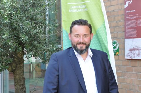 Ein Mann steht vor einer Flagge mit der Aufschrift "DVGW". Er lächelt freundlich und ist professionell gekleidet.