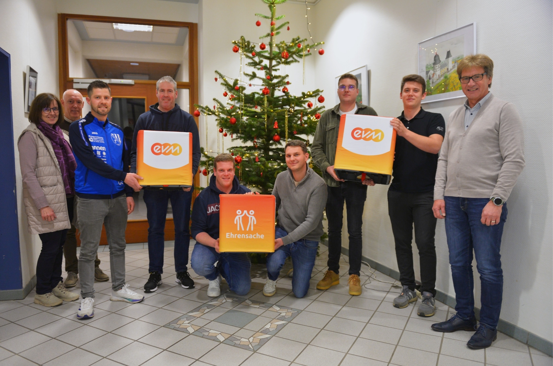Gruppenfoto im Foyer vor einem geschmückten Weihnachtsbaum: Vertreter von drei Sportvereinen aus der Verbandsgemeinde Kelberg erhalten im Rahmen der evm‑Ehrensache eine Spende von insgesamt 1.500 Euro und halten dazu Geschenkboxen mit evm‑ und Ehrensache‑Logo.