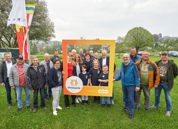 Eine Gruppe lächelnder Menschen steht draußen auf einer Wiese. Einige halten einen großen, orangefarbenen Rahmen mit der Aufschrift "Ehrensache" und "evm". Im Hintergrund sind Bäume und Fahnen zu sehen.